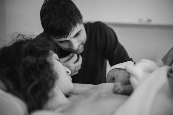 El papel de la pareja en el parto. Fotografía en blanco y negro de una madre sosteniendo a su bebé recién nacido en su pecho mediante contacto piel con piel, mientras su pareja se inclina con ternura para mirarlos de cerca en el hospital.